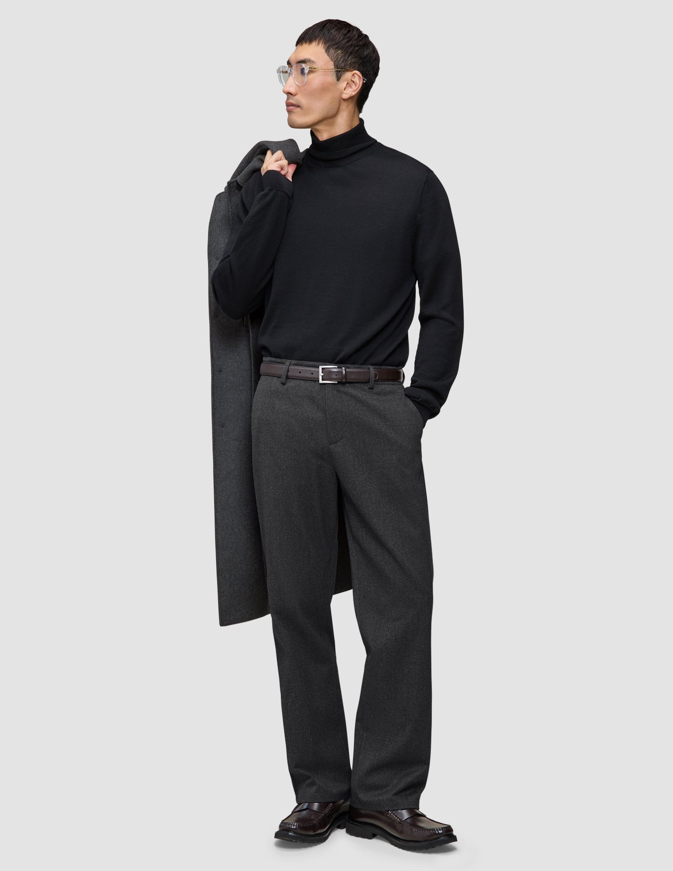 En mand i en sort rullekravebluse og mørkegrå Atelier Pants Relaxed, der har en frakke over skulderen.